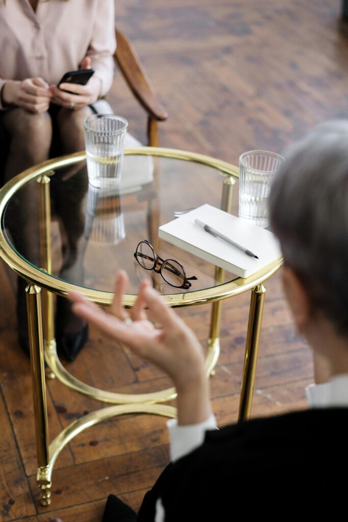 pexels-photo-4098374-4098374 Two adults engaged in a therapy session, seated at a glass table with notebooks and water glasses.